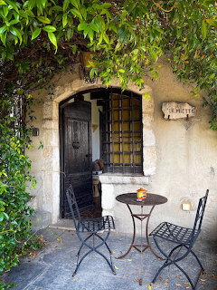 Bistro table and folding chairs under ivy-covered archway with rustic wooden doors in French courtyard