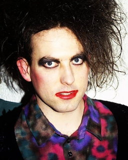 Robert Smith of The Cure with mesy black hair, dark eye makeup, red lipstick, and colorful shirt