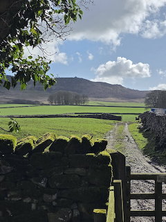Rural path beside stone walls and green fields with a hill under a cloudy blue sky
