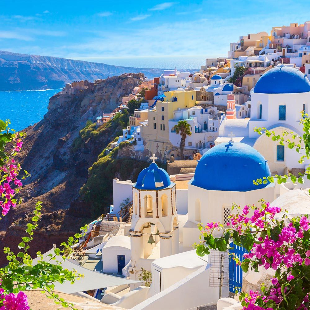 White buildings and blue-domed churches on cliff overlooking blue sea in Santorini with pink flowers