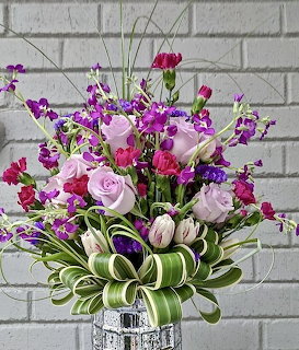 Purple and pink rose bouquet with orchids and green striped leaves in silver vase on white brick background