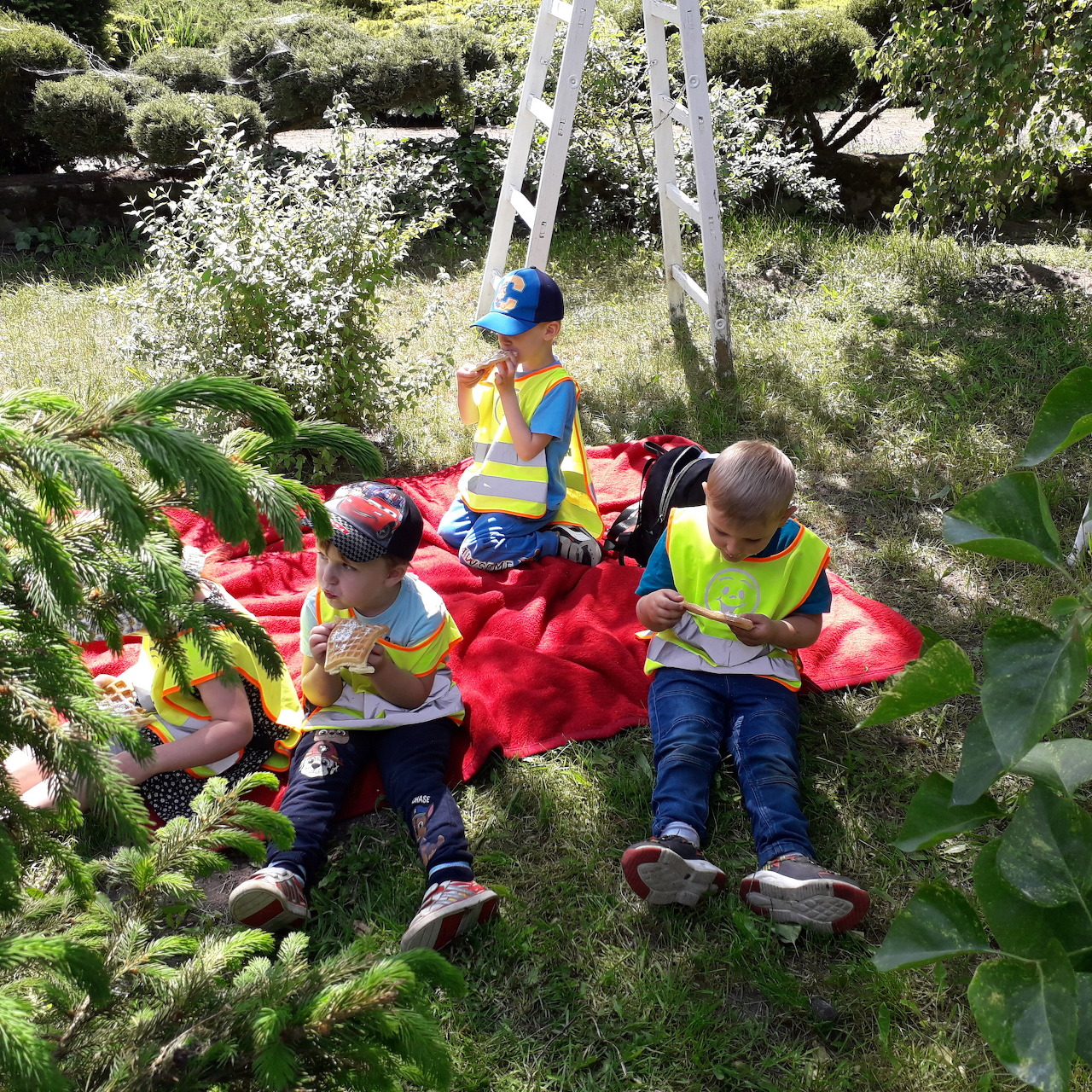 Three children in safety vests sitting on a red blanket outdoors in a garden