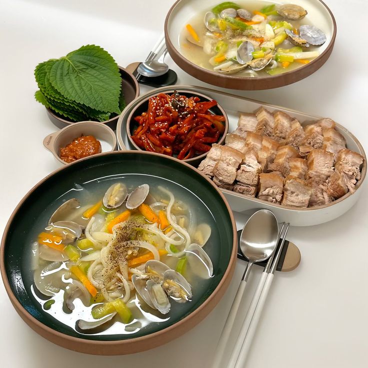 Overhead view of Korean food dishes with clam soup, spicy side dish, sliced pork, perilla leaves, and sauce