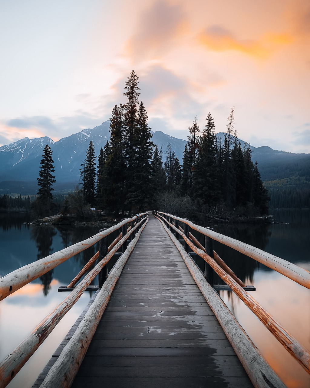 Wooden footbridge over a calm lake leading to pine trees with snow-capped mountains and sunset sky