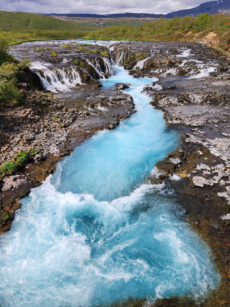 Bright turquoise glacial river and waterfall cascading over dark volcanic rocks in Iceland