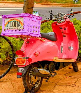 Pink scooter with purple Aloha crate parked on a street with tropical greenery in the background