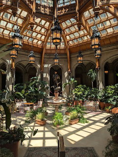 Elegant glass conservatory with wooden arched ceiling, hanging lanterns, and tropical plants surrounding a central fountain