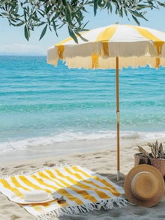 Yellow and white striped beach umbrella with fringed edges over a matching towel on a sandy beach by turquoise sea
