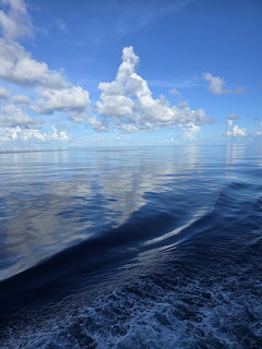 Blue ocean with a boat wake in the foreground and tall white clouds on the horizon