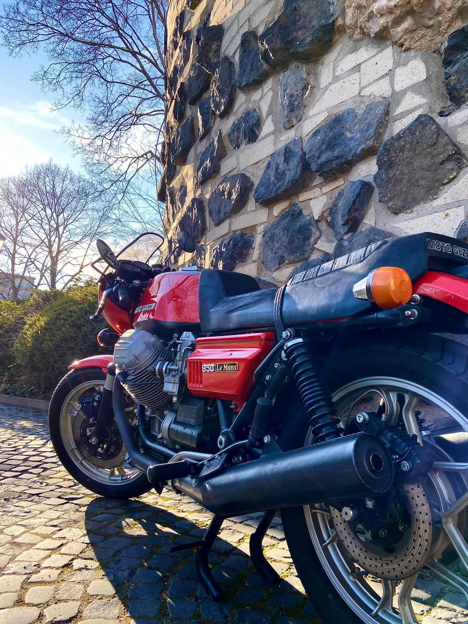 Red Moto Guzzi Le Mans MK1 vintage motorcycle parked beside a stone wall in sunlight
