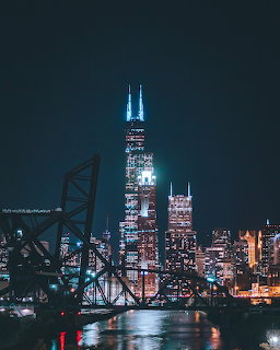 Night city skyline with lit skyscrapers, waterfront reflections, and industrial structures in the foreground.