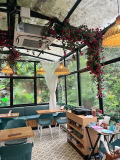 Glass-rofed cafe interior with red bougainvillea vines, woven lamps, teal chairs, and tropical greenery outside