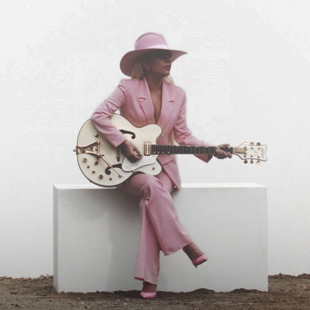 Woman in a pink suit and hat sitting on a white block with a white electric guitar