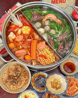 Divided hot pot filled with shrimp, crab, meat, vegetables, noodles, and side dishes on a red table