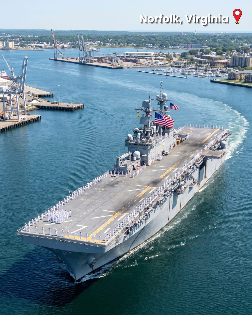 Large gray naval ship moving through a harbor with docks and city buildings in the background