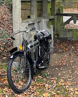 Vintage black AJS motorcycle beside a wooden gate with fallen leaves on the ground