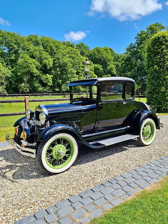 Black 1929 Ford Model A vintage car with green wire wheels on gravel driveway with trees