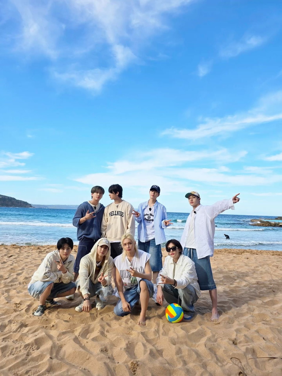 Group of seven young men posing on a sunny beach with ocean waves and blue sky behind them.