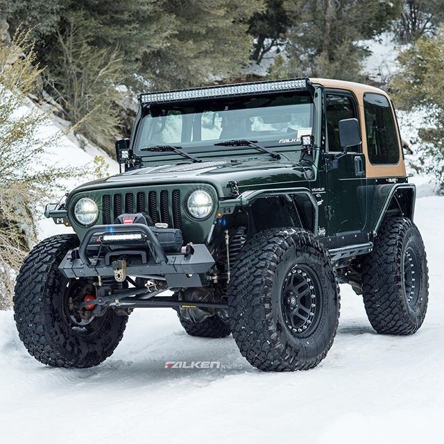 Modified green Jeep Wrangler with large off-road tires parked in snow with pine trees in background