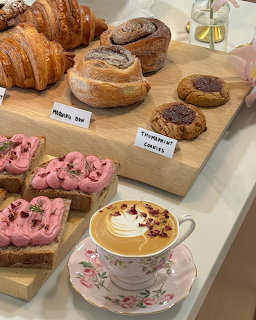 Wooden board with croissants, cinnamon rolls, thumbprint cookies, pink frosted cake slices, and a floral latte cup