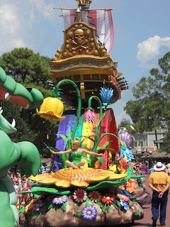 Disney parade float with Tinkerbell figure, golden ornate tower, and costumed characters in outdoor theme park setting
