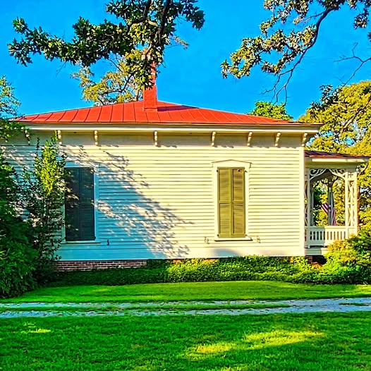 White wooden house with red roof, green lawn, and trees in bright sunlight