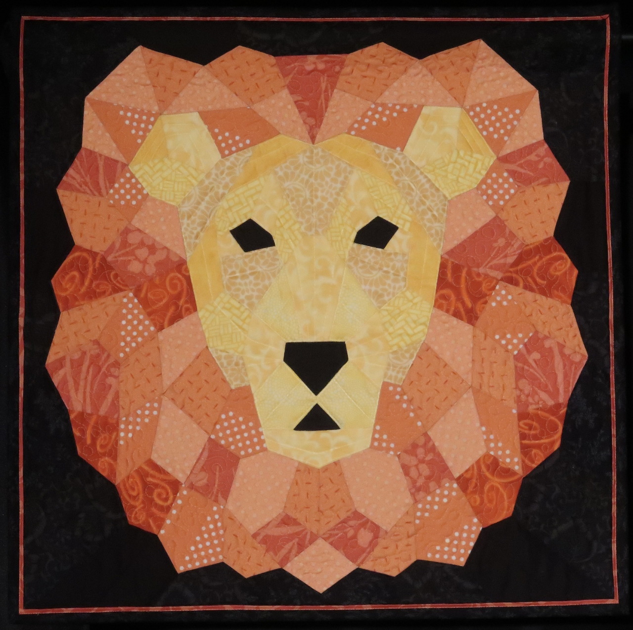 Patchwork quilt showing a stylized lion face made of orange and yellow geometric fabric pieces on black background