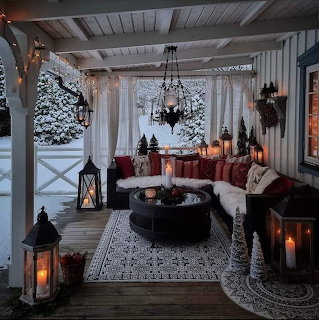 Cozy covered porch with white sofa, lanterns, candles, and snowy evergreens outside during winter evening