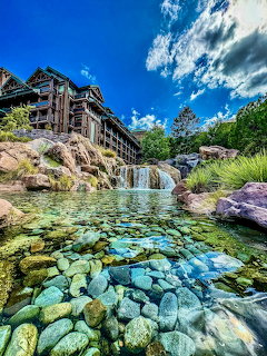 Rustic wooden lodge building with waterfall cascading into clear pool filled with colorful rocks under blue sky