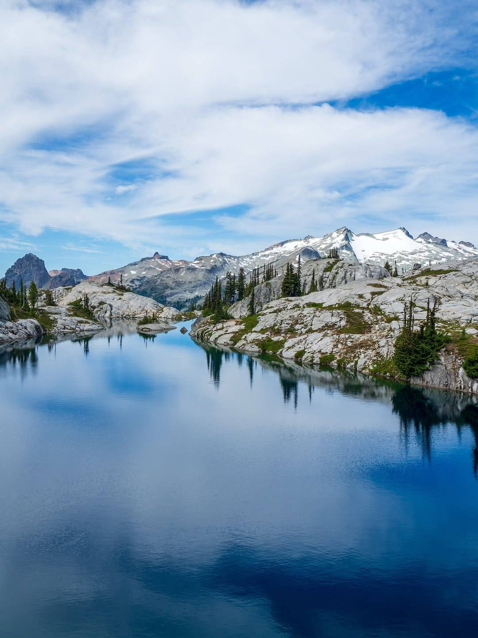 Calm alpine lake reflecting snow-capped mountains and rocky terrain under a blue sky with clouds