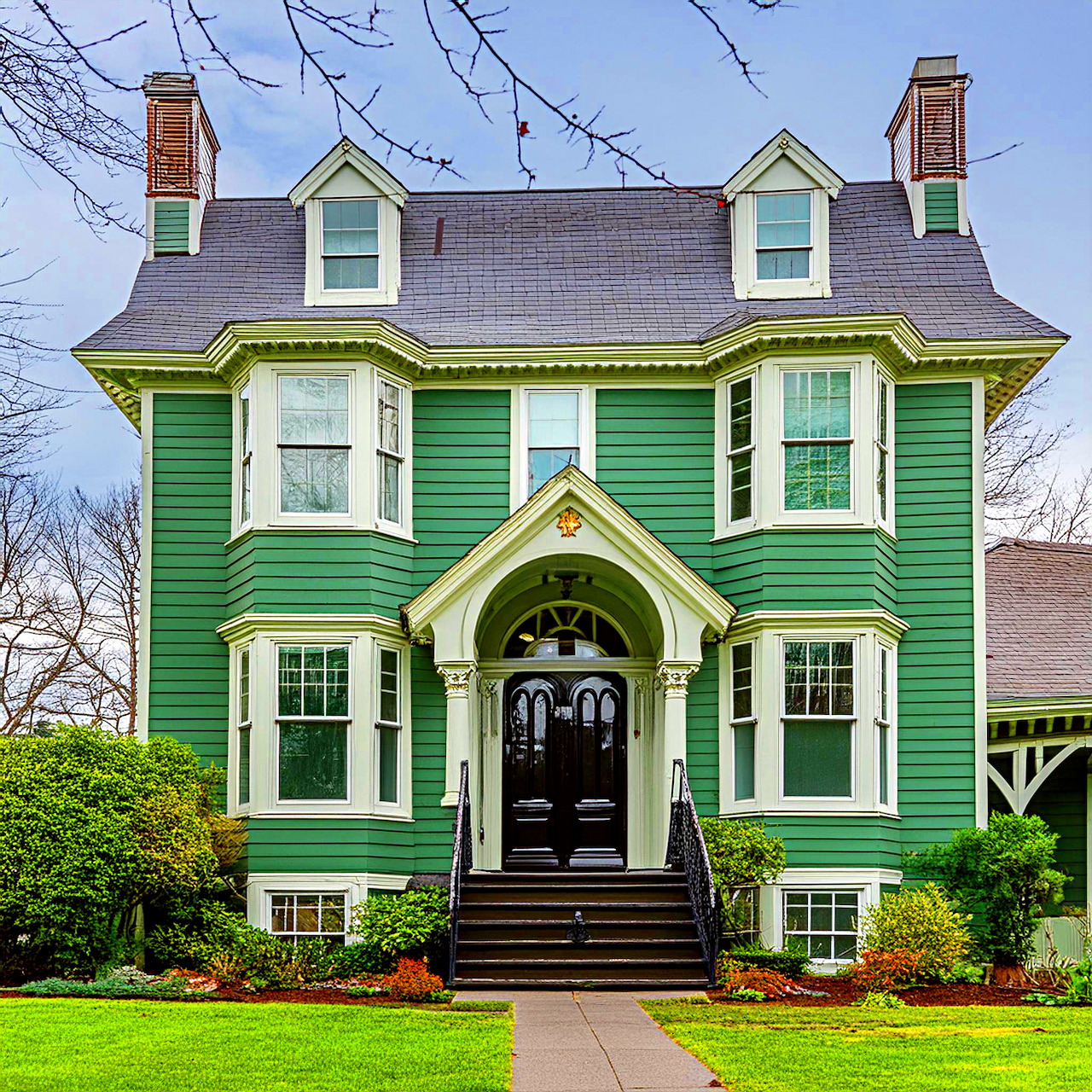 Green Victorian house with white trim, bay windows, and black front door surrounded by lawn and trees