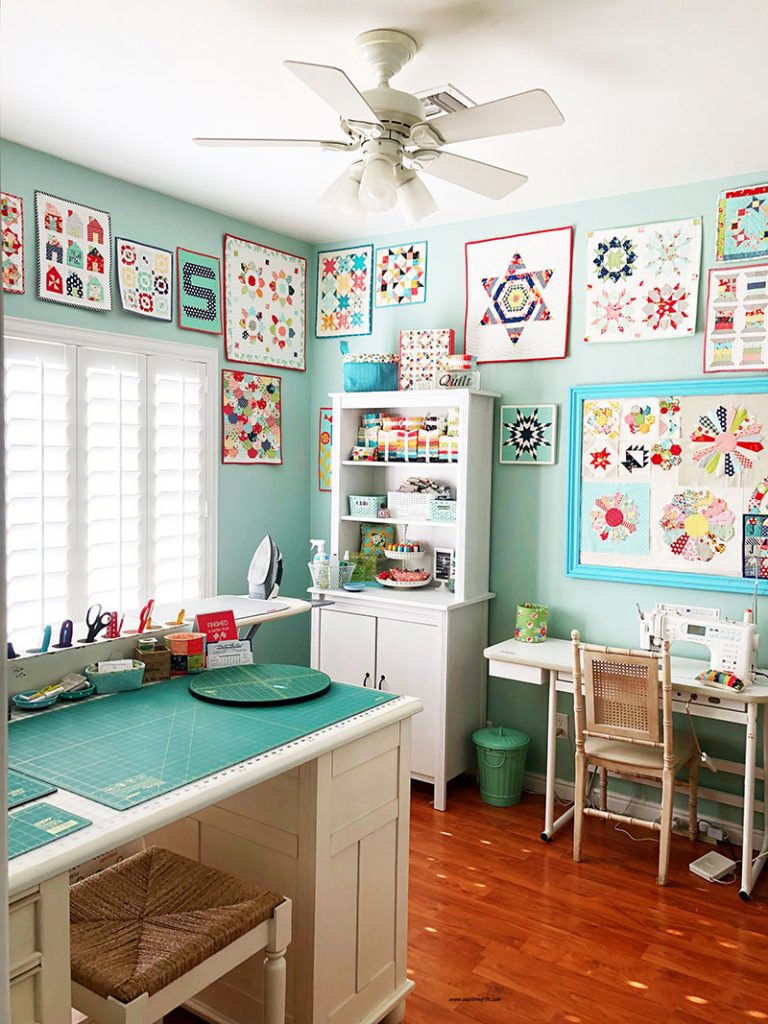 Cheerful sewing room with turquoise table, wooden desk, shelves, and colorful quilt art on mint green walls