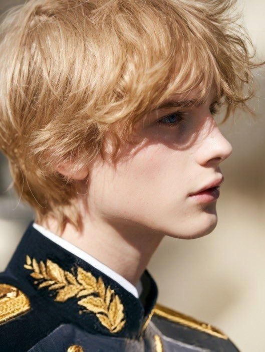 Side-profile portrait of a blond youth in a dark jacket with gold embroidered trim