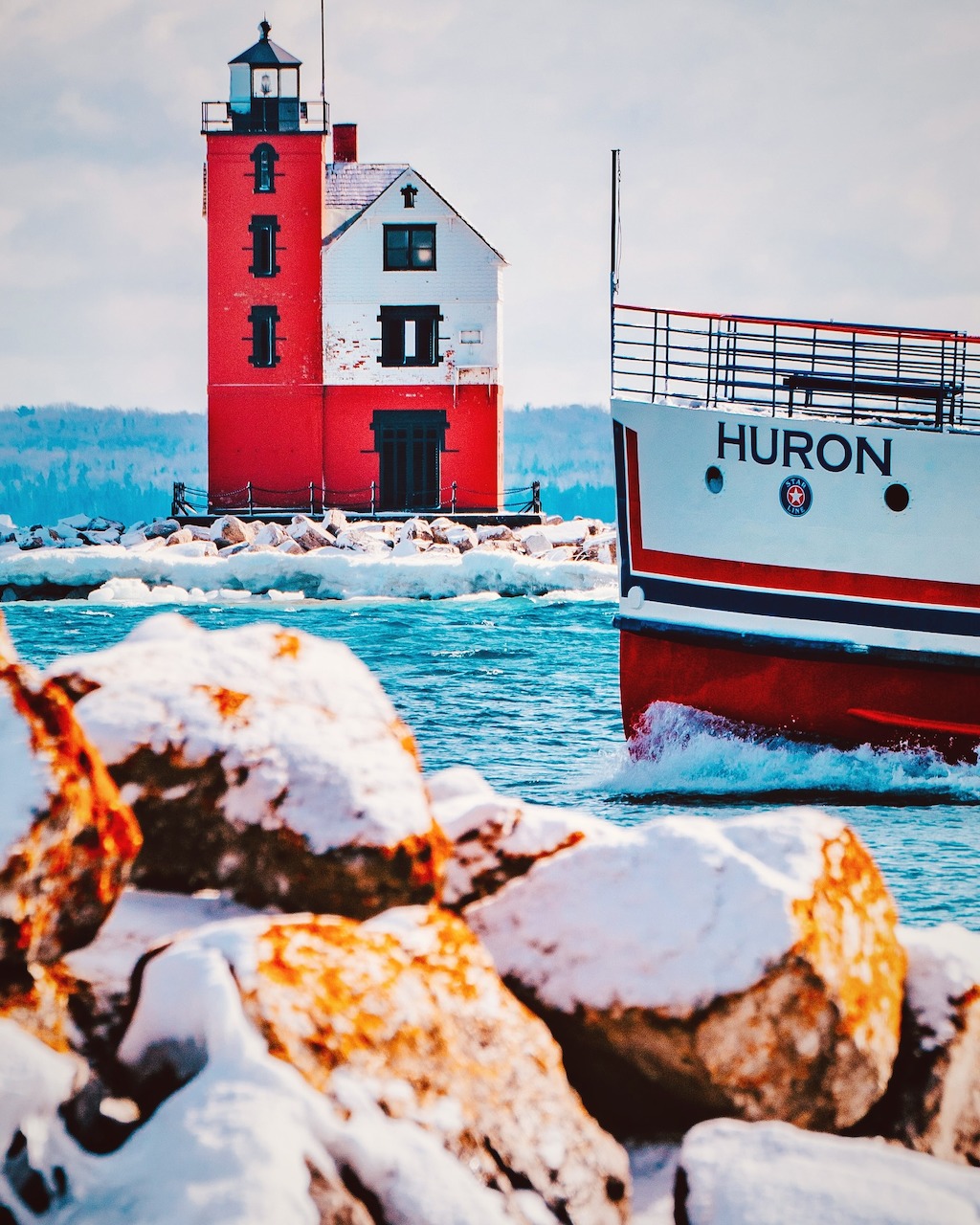 Red lighthouse on snowy breakwater next to boat named Huron in icy harbor