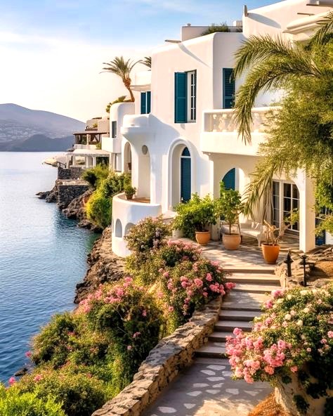 White Greek villas with teal shutters on a rocky seaside cliff with pink flowers and palm trees