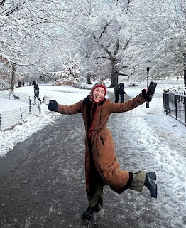 Woman in brown coat kneeling on snowy path with arms raised, surrounded by snow-covered trees in winter park