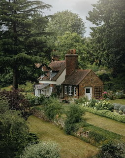 Brick English cottage surrounded by tall trees, green lawn, and flower gardens in a countryside setting