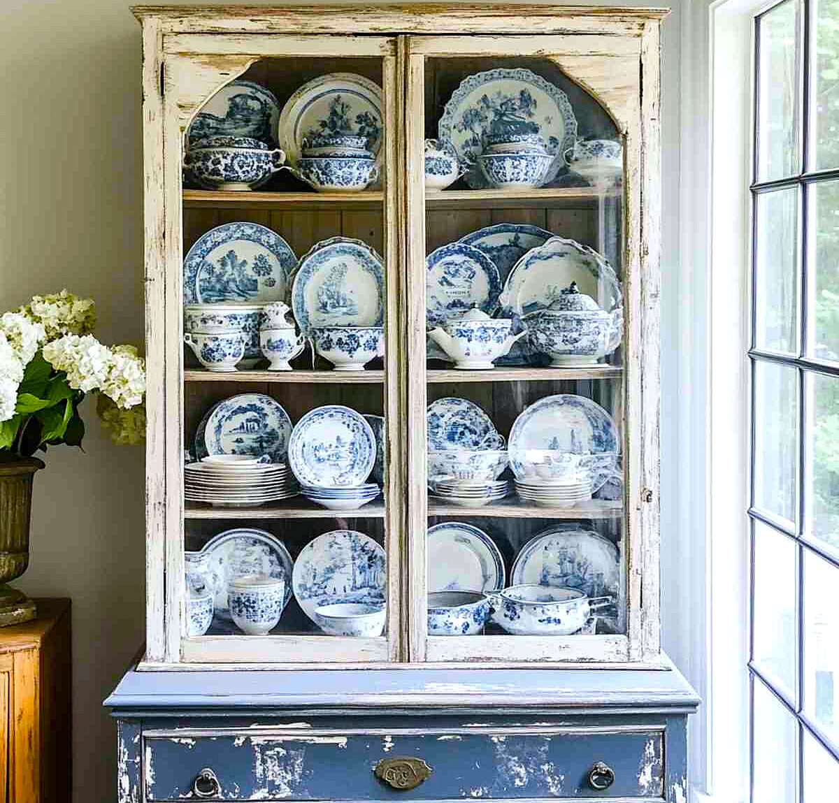 Distressed white cabinet filled with blue and white china plates and bowls behind glass doors