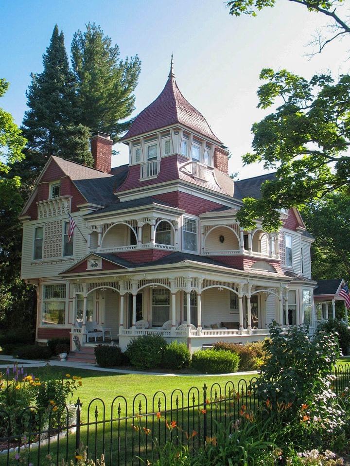 Large Victorian house with red turret, wraparound porch, and ornate white detailing amid green trees