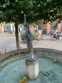 Bronze statue fountain in a coblestone town square shaded by green trees
