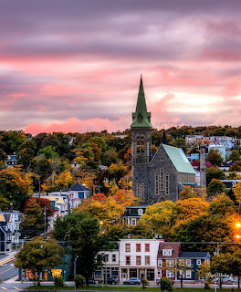 Stone church with tall spire surrounded by autumn trees and colorful houses under a pink sunset sky