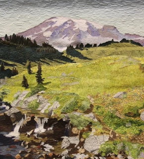 Art quilt showing a snow-capped mountain with green meadows, pine trees, and a rocky creek