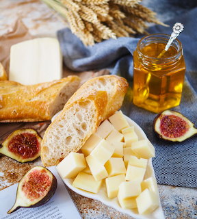Cheese board with cubed cheese, bread, figs, honey jar, and wheat on blue fabric