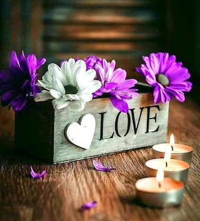 Wooden box displaying LOVE with white heart, purple and white daisies, and lit candles on wood table