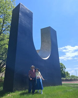 Two people posing beside a tall black U-shaped sculpture outdoors on grass under a blue sky.