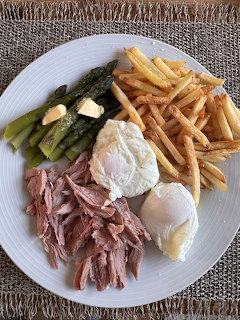 White plate with ham, fried egg, chips, asparagus, and a poached egg on a woven mat.