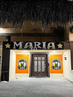Entrance of Maria Bar with sign, central wooden door, and orange panels beneath a thatched roof