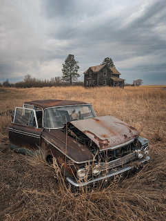Rusty abandoned AMC Rambler in a dry field with an old farmhouse in the background.