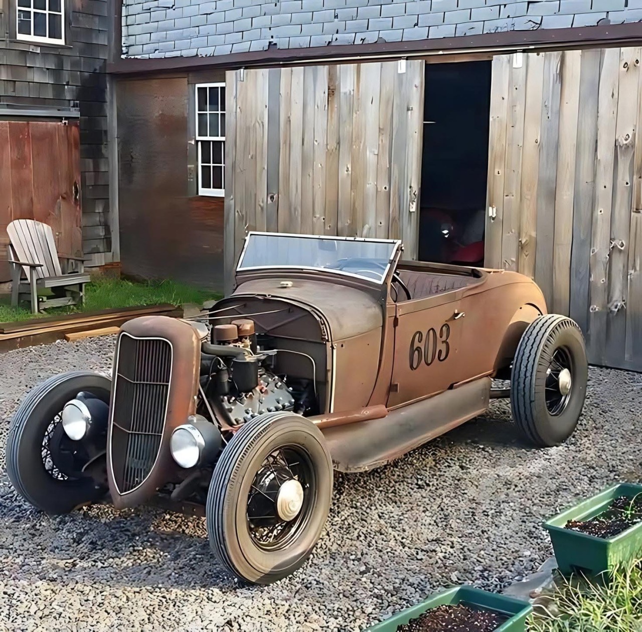 Rusty vintage hot rod with exposed engine parked on gravel by wooden sheds