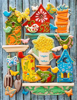 Colorful decorated sugar cookies shaped like birdhouses, rain boots, flowers, birds, and garden tools arranged on a wooden shelf backdrop.